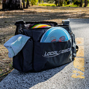 Local Discs disc golf bag with discs, rag and bottle on a tee pad